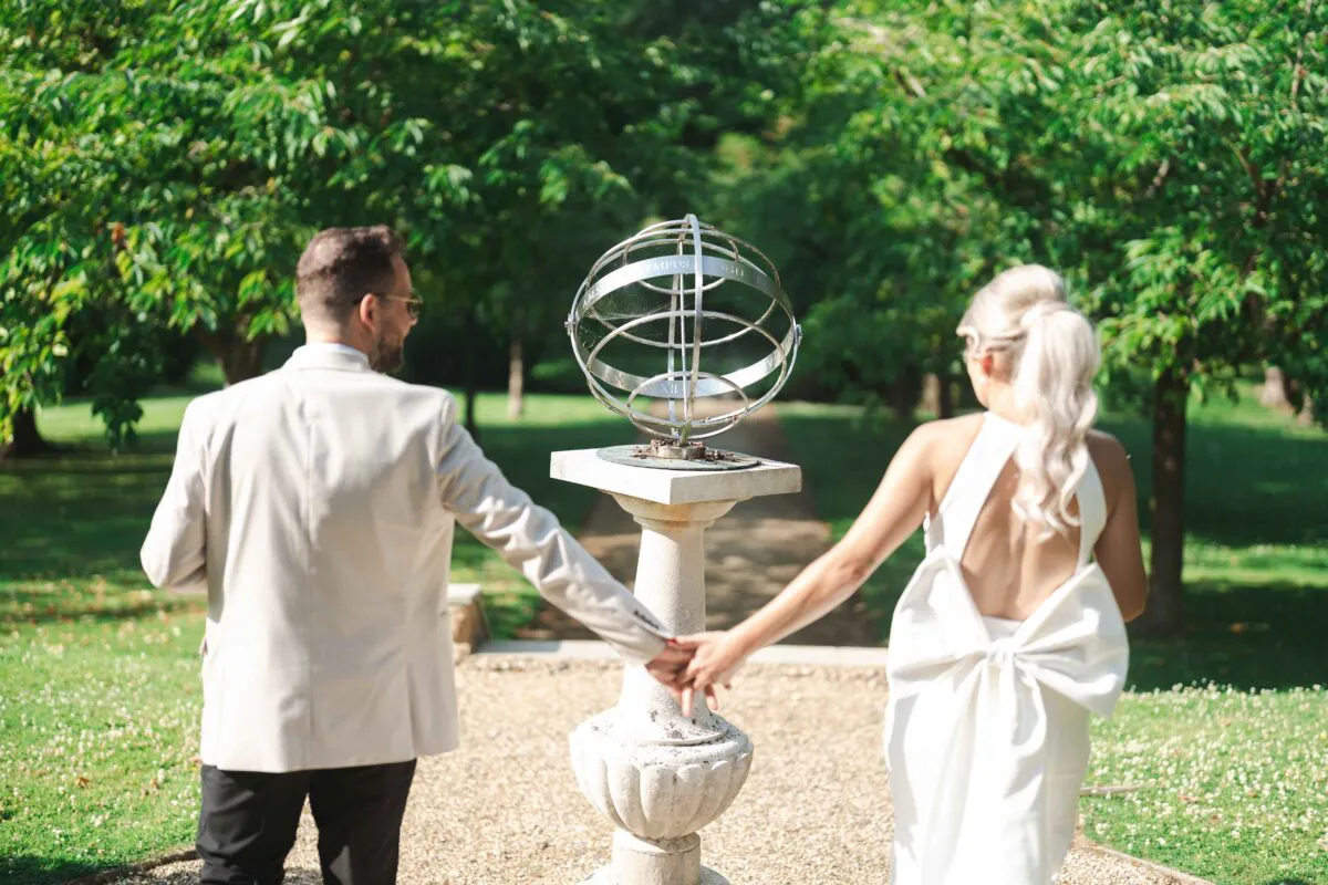 Newlyweds walk hand in hand away from the camera through a sun-dappled garden past an ornate stone sundial.