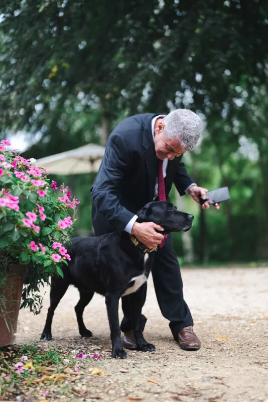 A guest in a black suit lovingly interacts with a black dog outdoors near a planter filled with vibrant pink flowers.