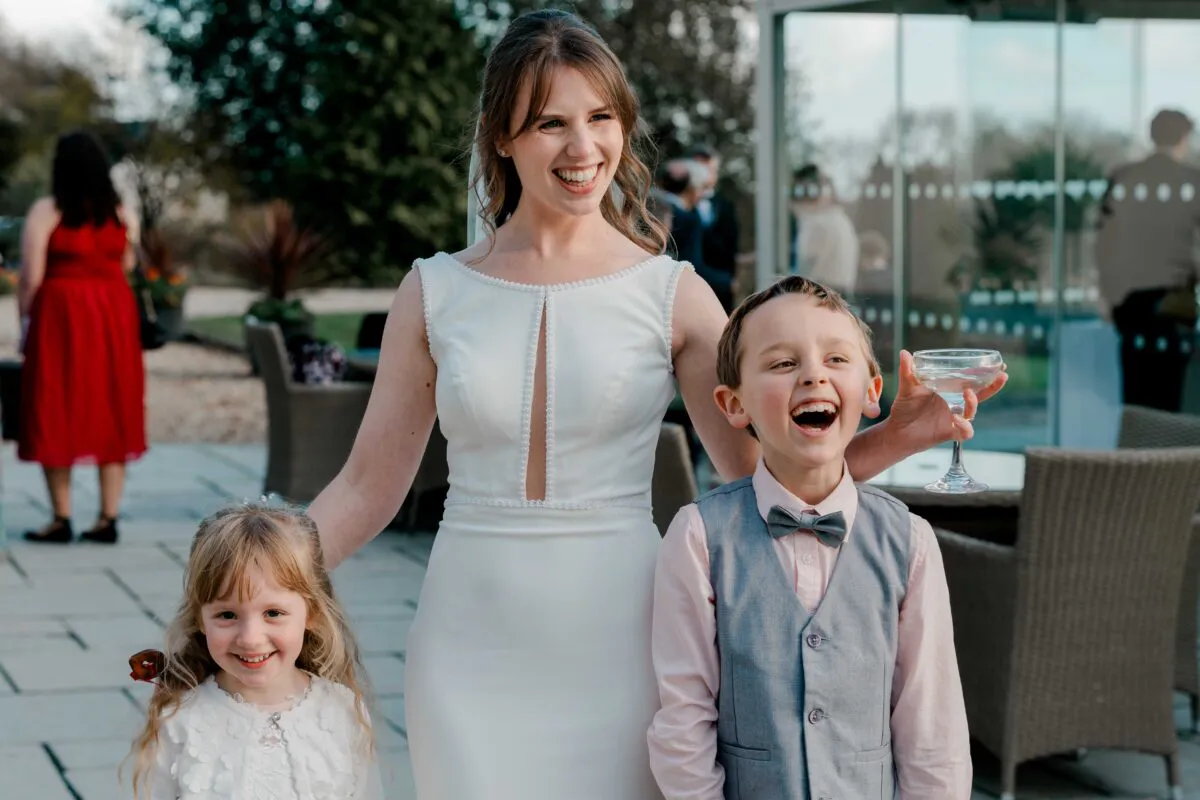 A joyful bride laughs alongside a young page boy in a grey waistcoat and a smiling flower girl during an outdoor drinks reception.