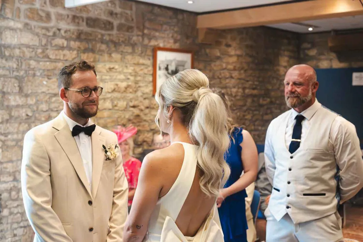 A groom in a beige suit with a black bow tie looks lovingly at his bride, who wears an open-backed gown with a high ponytail, during their indoor ceremony against an exposed stone wall.