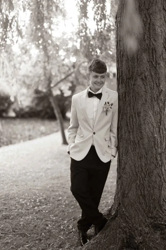 A young page boy wearing a suit and bow tie leans against a large tree trunk in a woodland setting during an outdoor wedding.