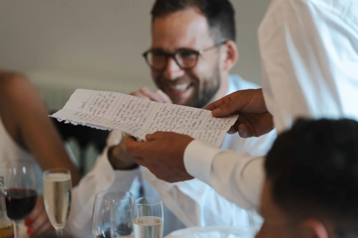A smiling man holding handwritten speech notes while guests laugh during the wedding reception.