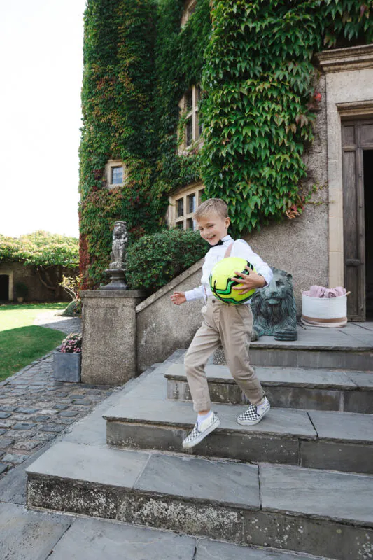 A young page boy happily carrying a bright football while bounding down the stone steps of an ivy-covered country house venue.