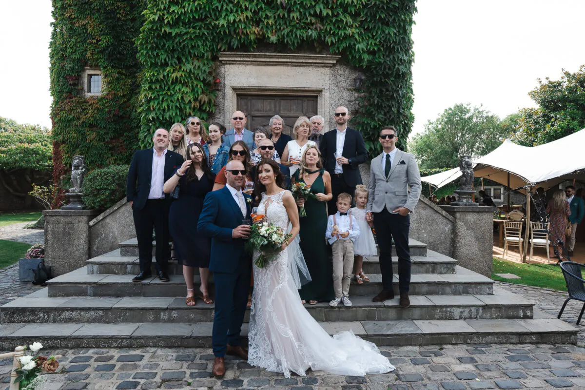 The bride and groom pose with family and friends on grand stone steps in front of an ivy-covered historic building during a summer wedding celebration.
