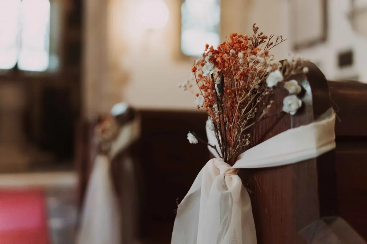 A wooden church pew decorated with a cluster of dried orange flowers and white blossoms secured with a delicate white ribbon.