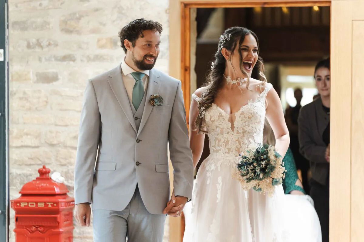A newlywed couple walk hand in hand through a wooden doorway, laughing joyfully as they enter a rustic stone venue.