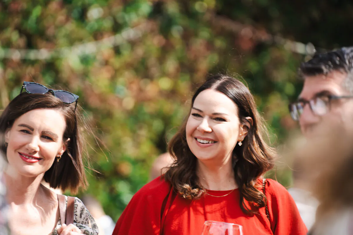 Smiling wedding guests mingle outdoors in the sunshine, with two women laughing candidly amidst natural foliage.