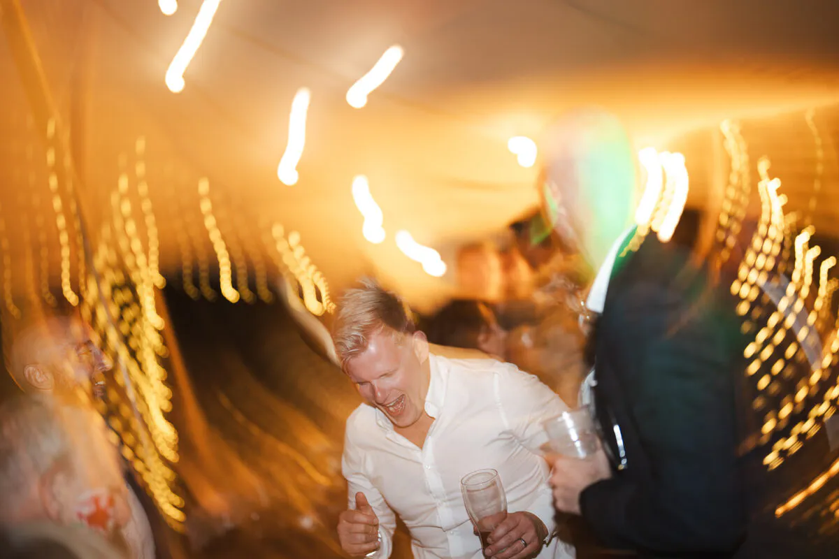 Guests enjoy a lively wedding reception under a marquee with dynamic motion blur and warm string lighting overhead.