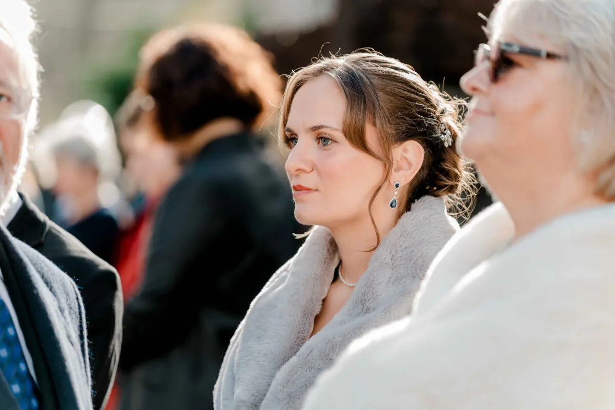 A candid outdoor shot captures a wedding guest wearing a faux fur stole and drop earrings, looking intently during the ceremony.