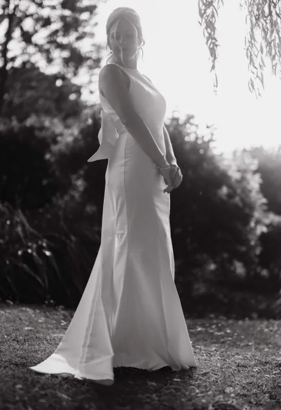 A stunning black and white portrait of a bride in a high neck crepe gown standing outdoors with gentle backlighting.