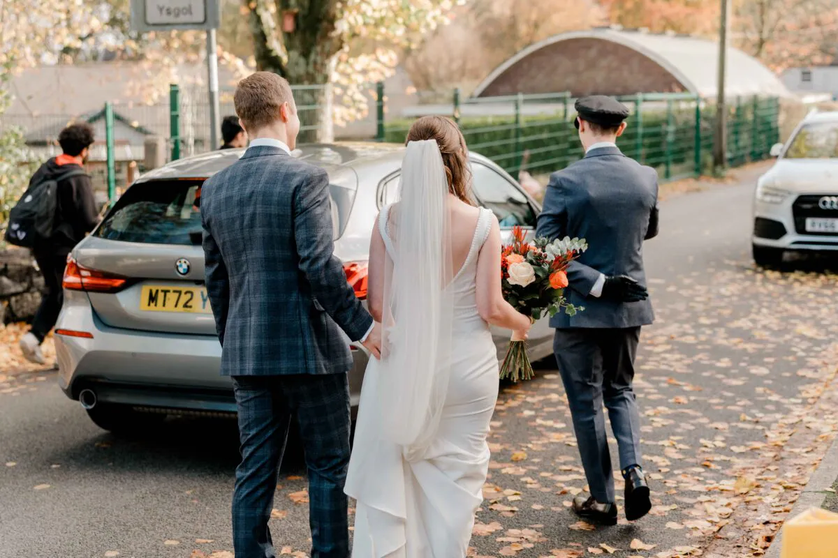 A newlywed couple walks hand in hand along a leaf-strewn street towards a wedding car, accompanied by a chauffeur.