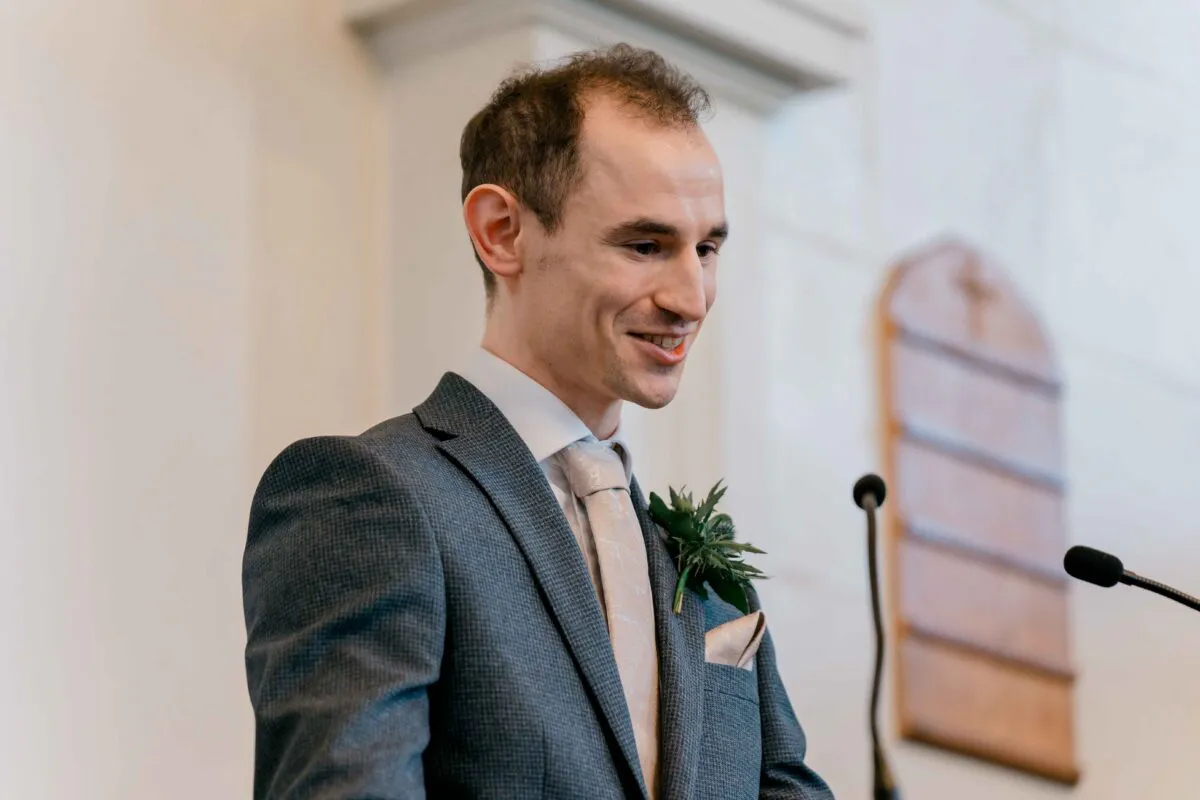 A smiling groom delivers his speech while wearing a grey suit, patterned tie, and a thistle buttonhole during the wedding reception.