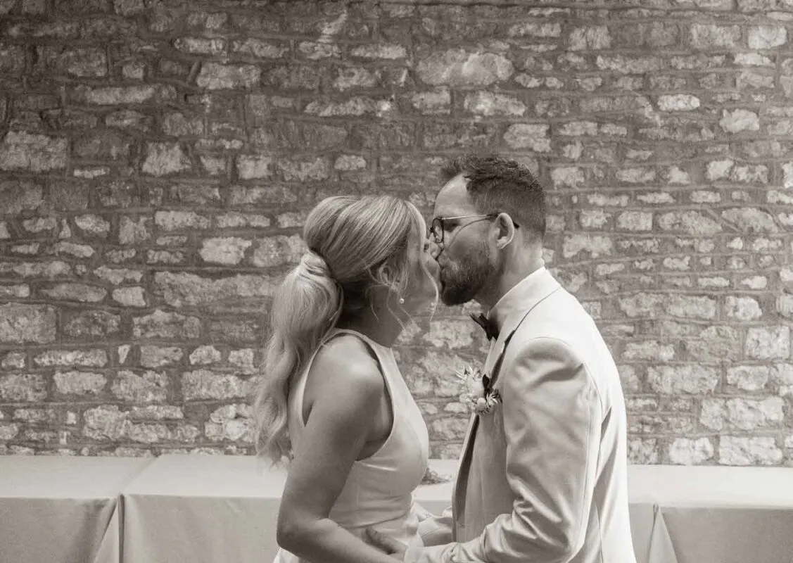 A newlywed couple share a tender kiss in front of a rustic exposed stone wall inside a wedding venue.