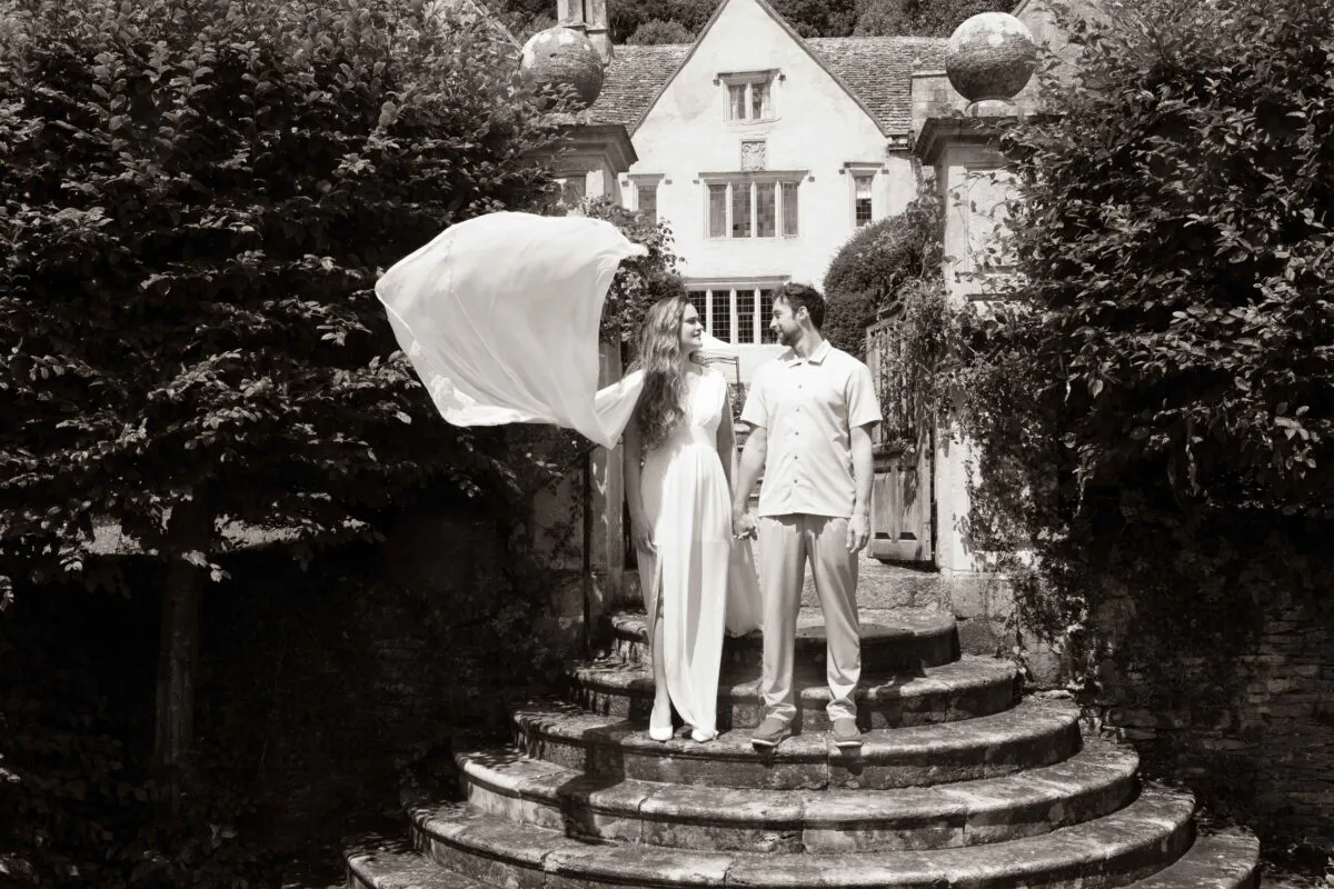 A newlywed couple holds hands on stone steps outside a historic country house as a light veil billows in the wind.