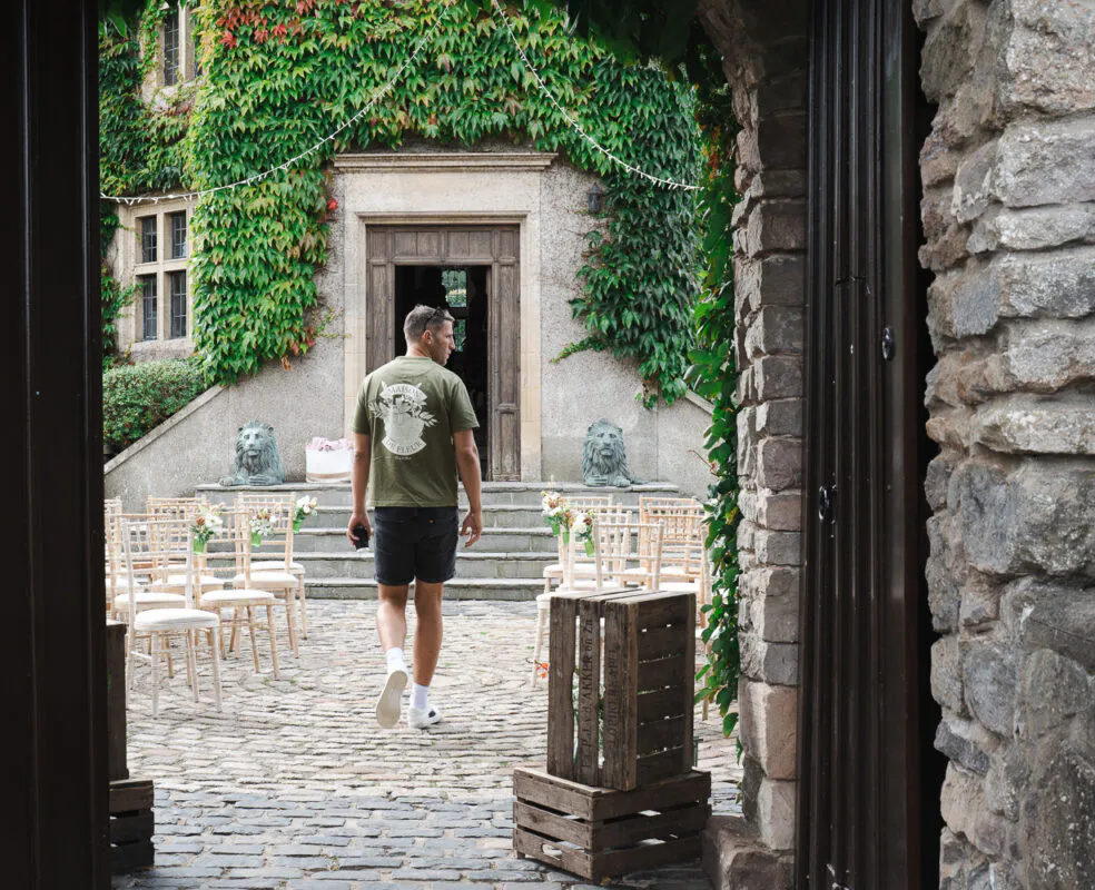 A man walks away across a cobbled courtyard towards an ivy-covered stone building set up for an outdoor wedding ceremony.