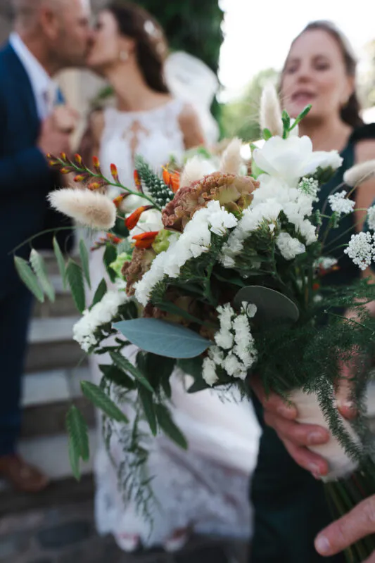A close-up shot of a textured wedding bouquet featuring white flowers, greenery, and dried grasses, with a blurred bride and groom kissing in the background.