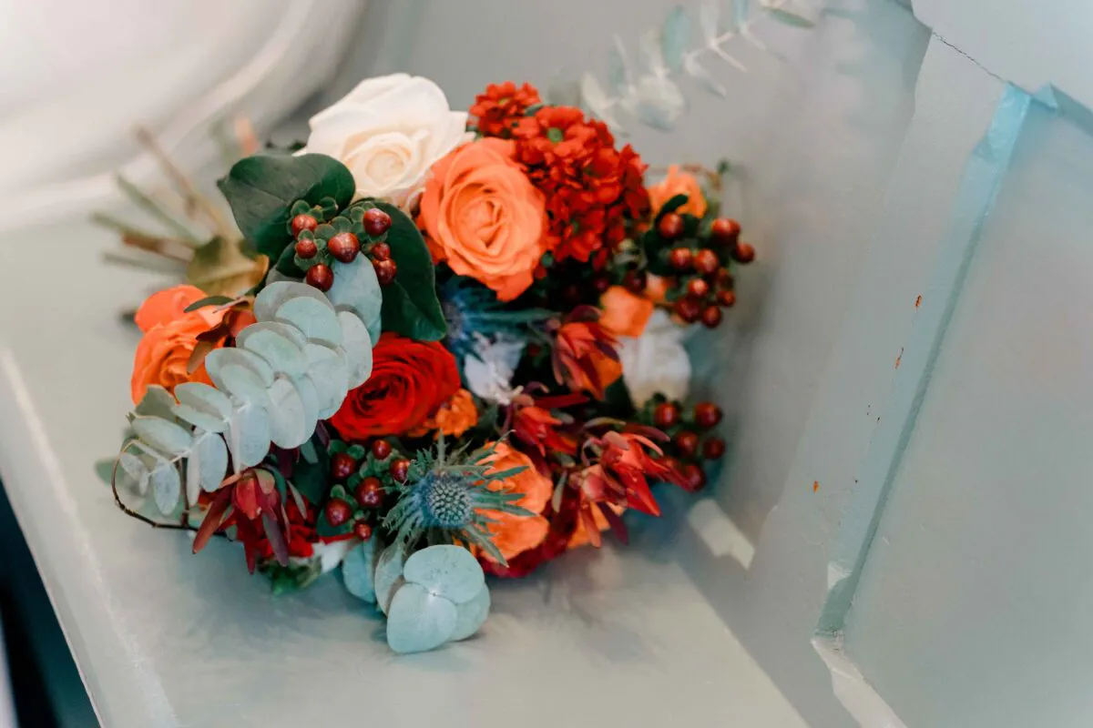 A vibrant hand-tied wedding bouquet featuring bright orange roses, deep red roses, white roses, thistles, and eucalyptus rests against a light blue panelled surface.
