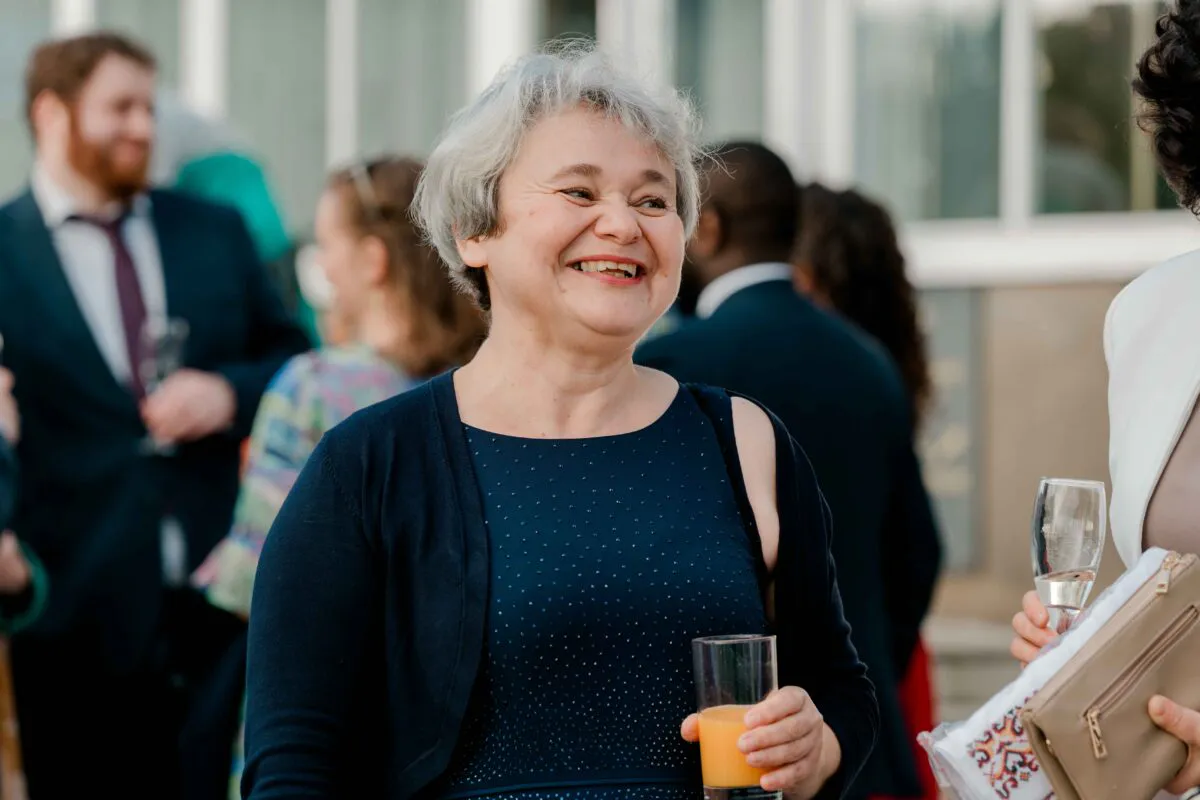 A mature guest with short grey hair laughs joyfully while holding a glass of juice during an outdoor drinks reception.