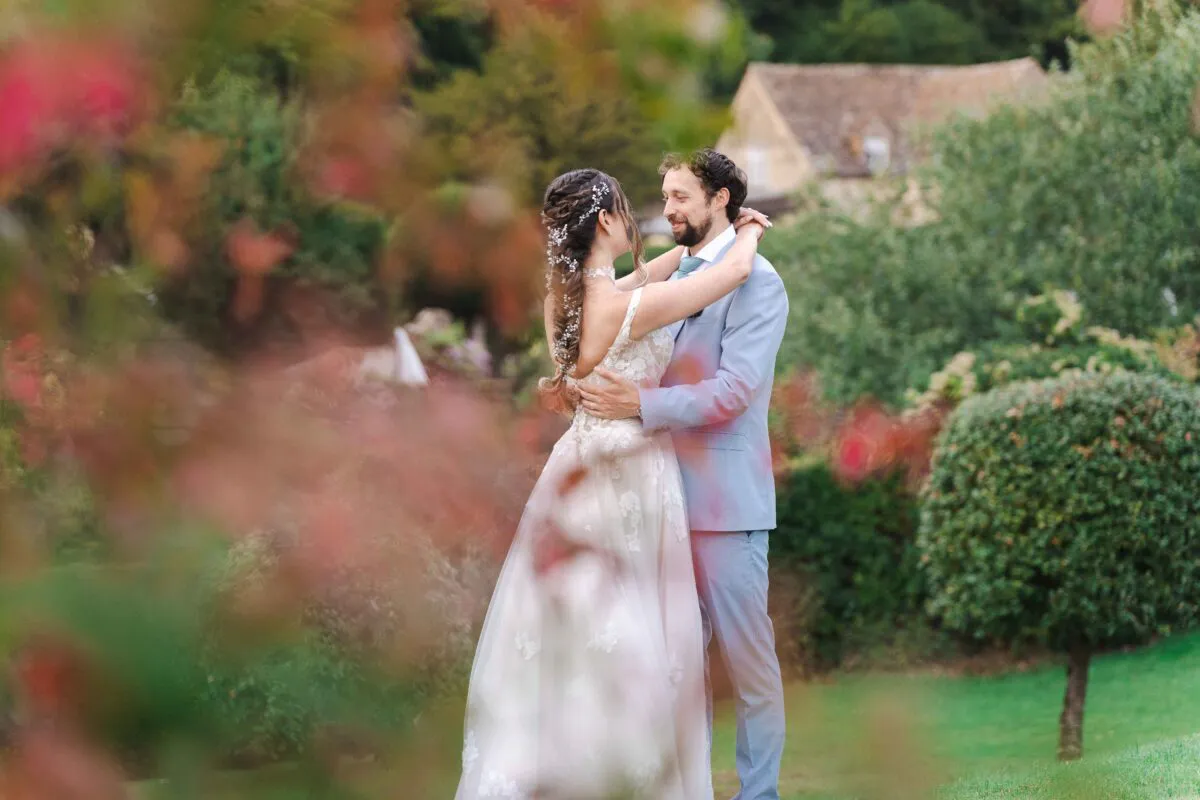 A newlywed couple embrace lovingly in an english country garden, framed by softly blurred foliage in the foreground.