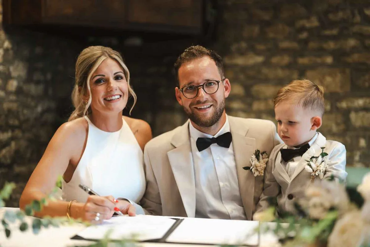 A smiling bride and groom sign the wedding register alongside a young page boy wearing a matching suit and bow tie.