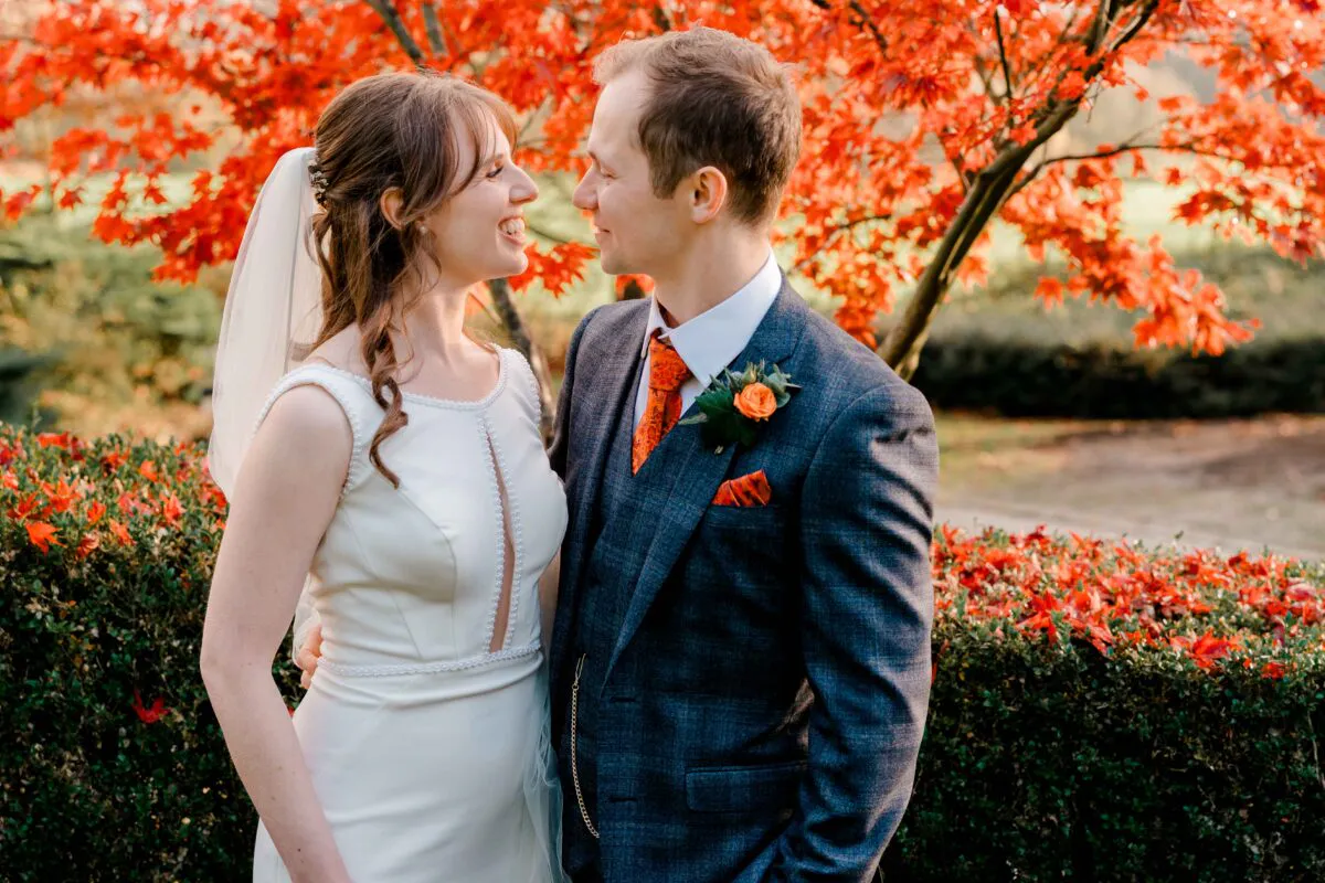 A smiling bride and groom stand outdoors against a backdrop of vibrant red autumn leaves during their wedding portrait session.
