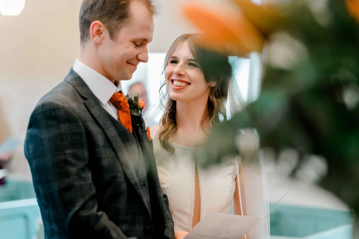 A smiling bride and groom share a joyful glance during their wedding ceremony, framed by soft foreground foliage and natural light.
