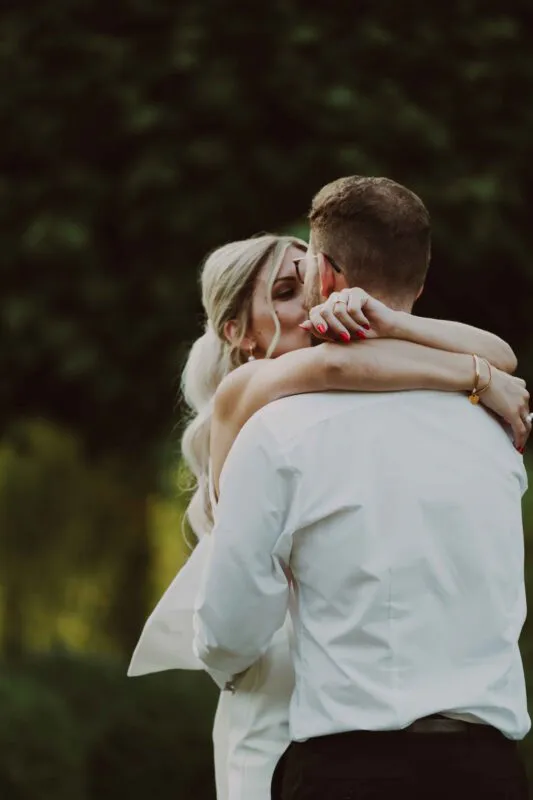 Newlyweds share an intimate embrace and kiss outdoors with soft sunlight filtering through the surrounding green foliage.