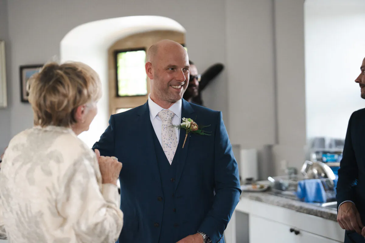 A smiling groom wearing a navy three-piece suit and a patterned tie chats with guests indoors during wedding celebrations.