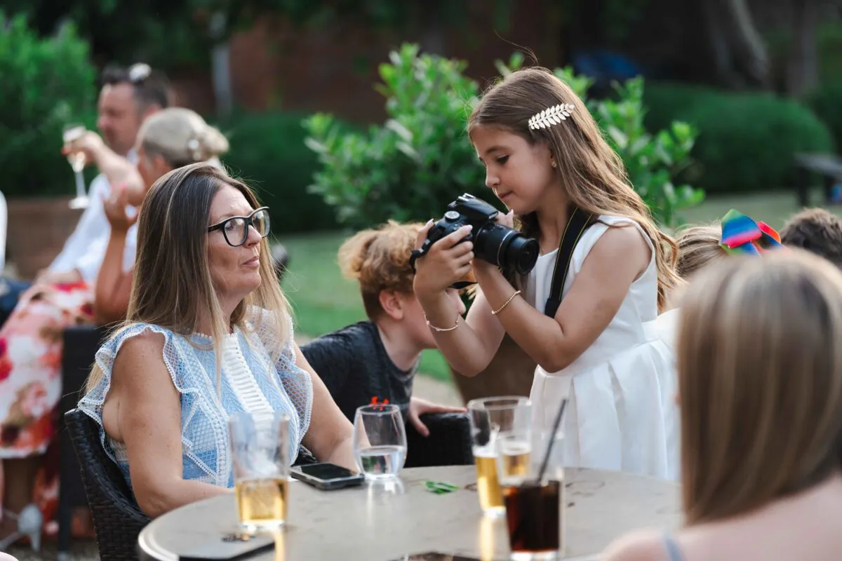 A young girl in a white dress holding a camera at an outdoor wedding reception table while guests mingle in the background.