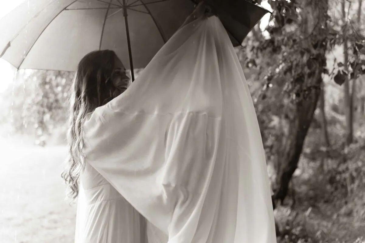 A smiling bride shares a joyful and intimate moment under a shared umbrella while draped beneath her wedding veil outdoors in the rain.