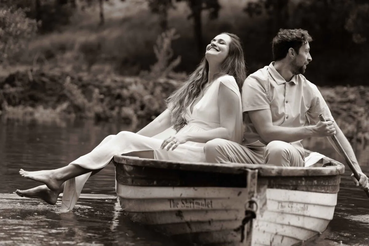 A newlywed couple laugh joyfully back-to-back in a wooden rowboat on a calm lake, with the bride trailing her bare feet in the water.