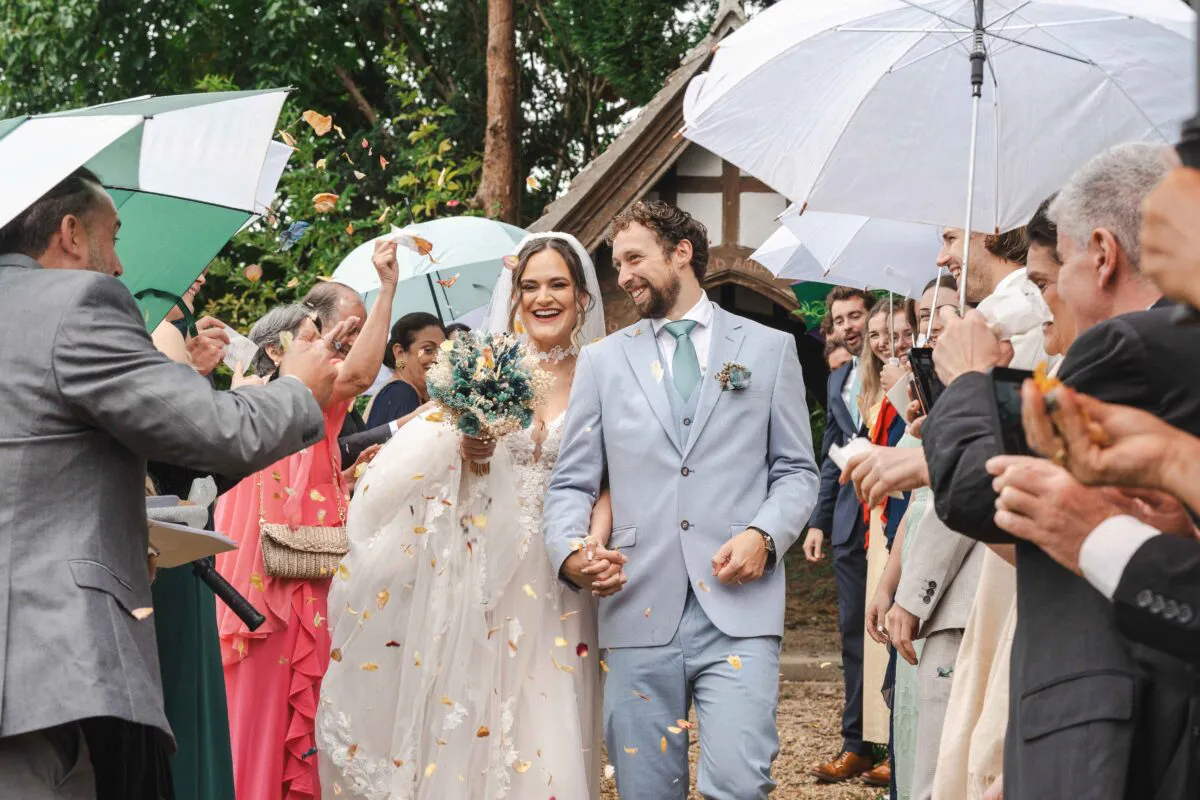 A joyful newlywed couple walks hand in hand through a shower of confetti outside a church on a rainy day, sheltered by white and green umbrellas held by cheering guests.