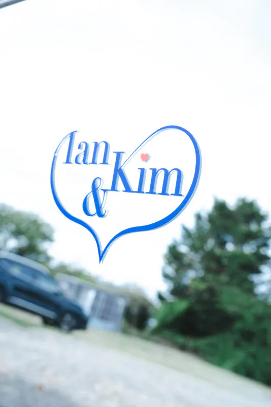 A personalised blue heart decal with the names Ian and Kim is affixed to a car window, with a softly blurred background of trees and a vehicle.