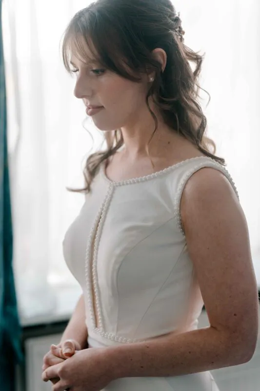 A beautiful bride stands by a window in natural light, showcasing her half-up hairstyle and a pearl-trimmed satin wedding gown during bridal preparations.
