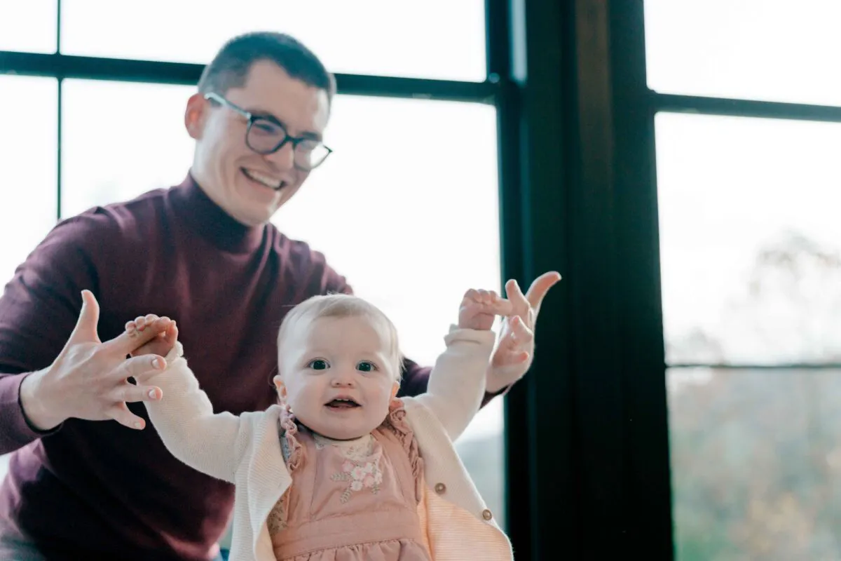 A smiling man holding a cheerful baby by the hands indoors near a large window.