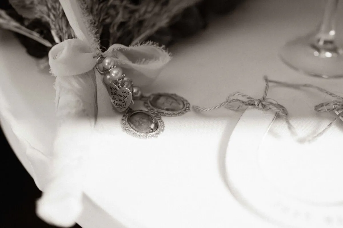 A close-up black and white shot captures a bridal bouquet stem adorned with a memorial charm holding small photographs and a heartfelt inscription.