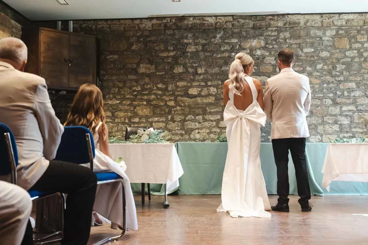 A bride and groom stand from behind facing an exposed stone wall during their indoor wedding ceremony, with guests seated on chairs in the foreground.