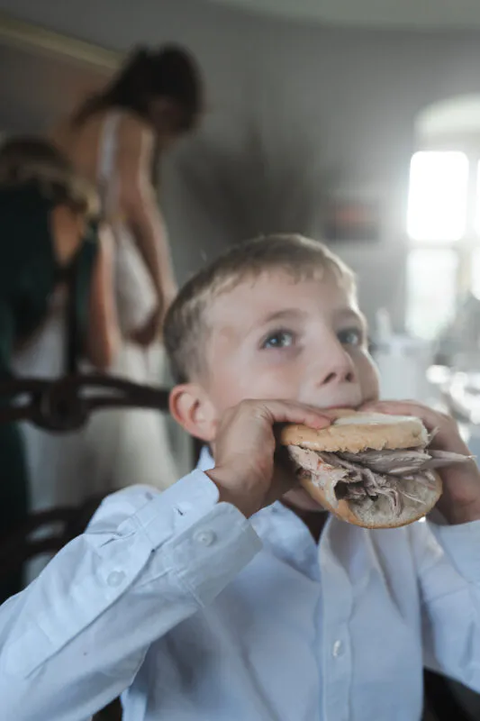 A young page boy in a white shirt enthusiastically takes a large bite out of a hog roast roll during the wedding reception.