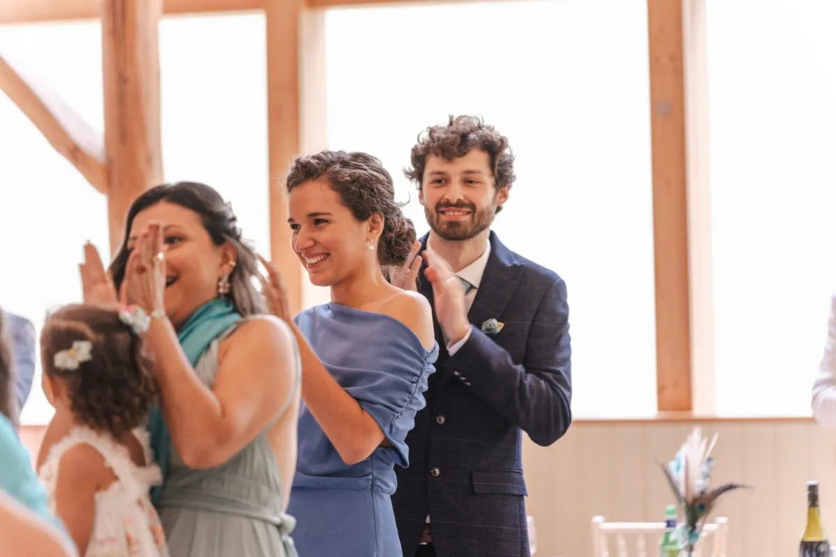 Wedding guests clapping and laughing joyfully inside a bright venue with large windows during the wedding reception.
