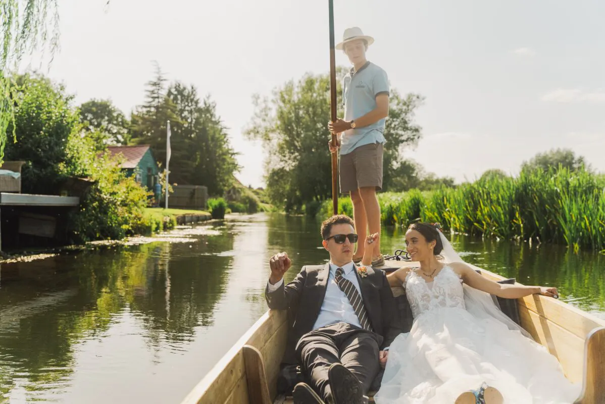 A newlywed couple relaxes in a wooden punt on a sunny river, accompanied by a chanter poling the boat through lush green countryside.