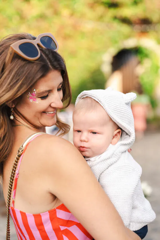 A smiling wedding guest wearing sunglasses on her head and floral face glitter holds a baby in a knitted grey hooded cardigan.