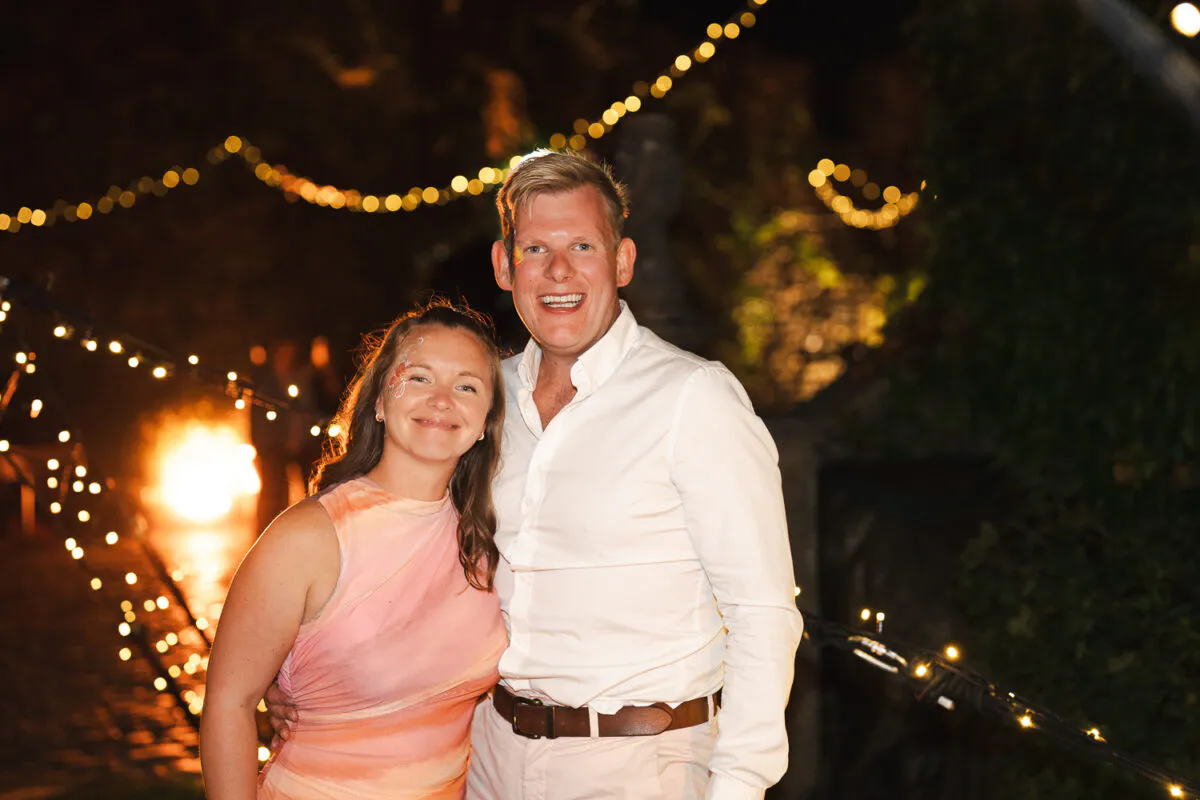 A smiling couple poses together outdoors at night under a warm canopy of fairy lights during an evening wedding celebration.