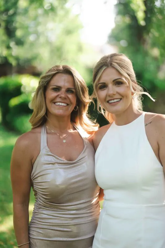 A smiling bride in a white halter neck dress stands outdoors next to her mother wearing a gold dress, both illuminated by soft natural light.