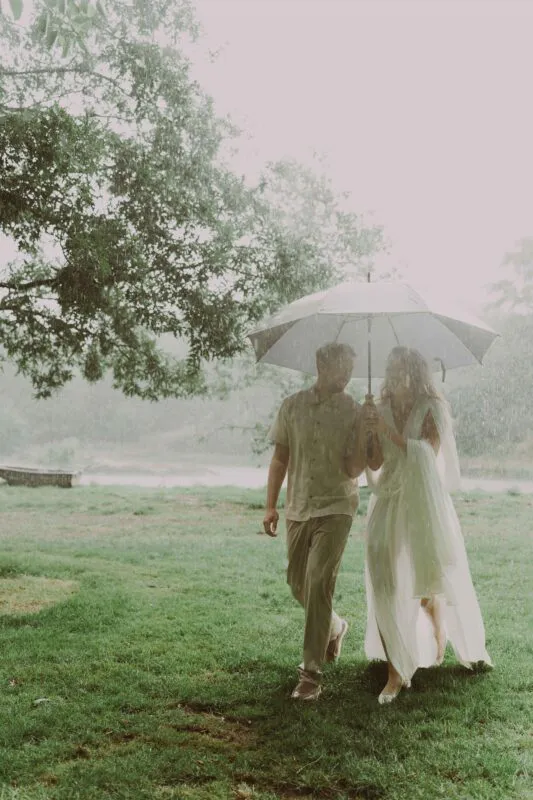 A bride and groom share a tender moment walking together under an umbrella during a gentle rain shower in a lush green lakeside setting.