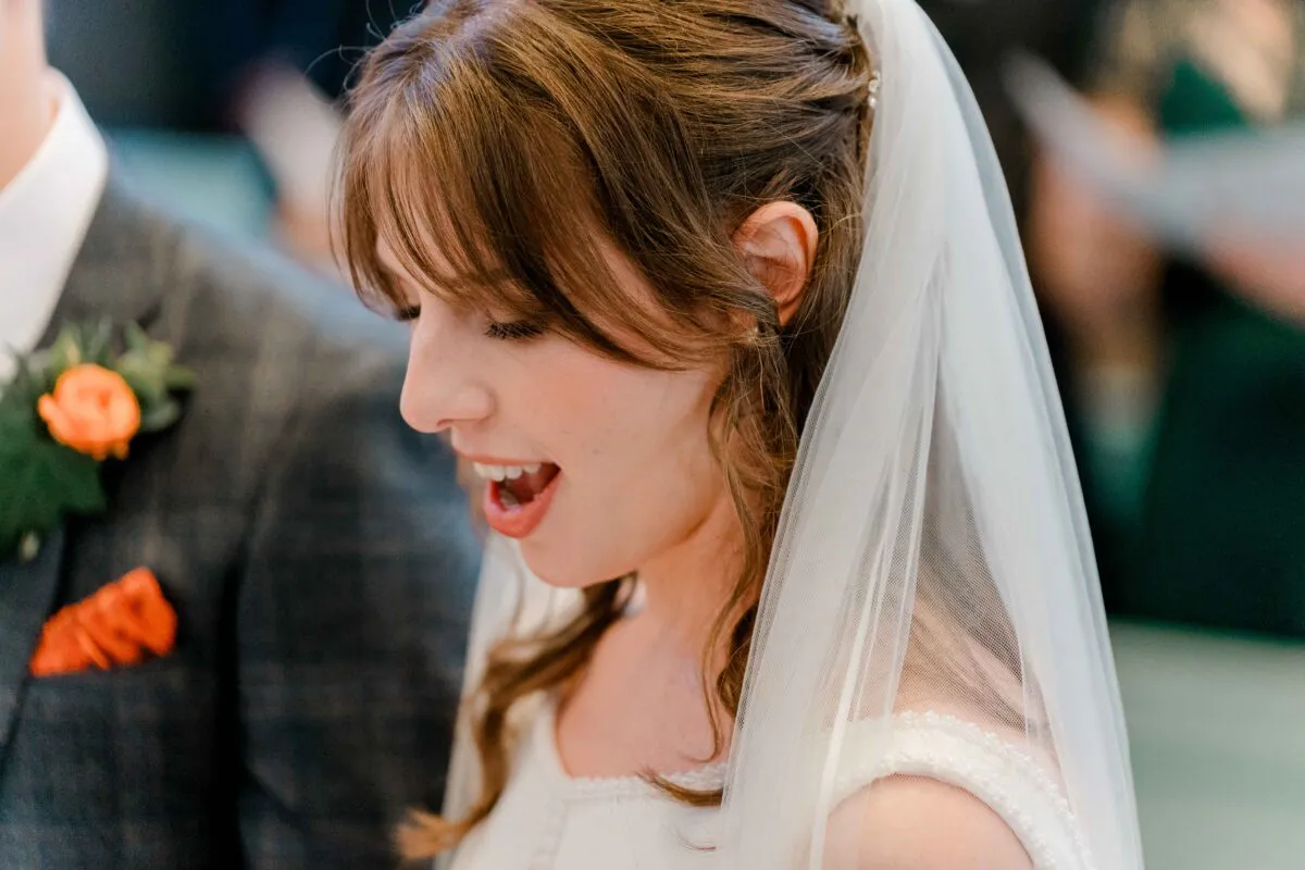 A smiling bride wearing a veil and a half-up hairstyle sings during the wedding ceremony alongside the groom.