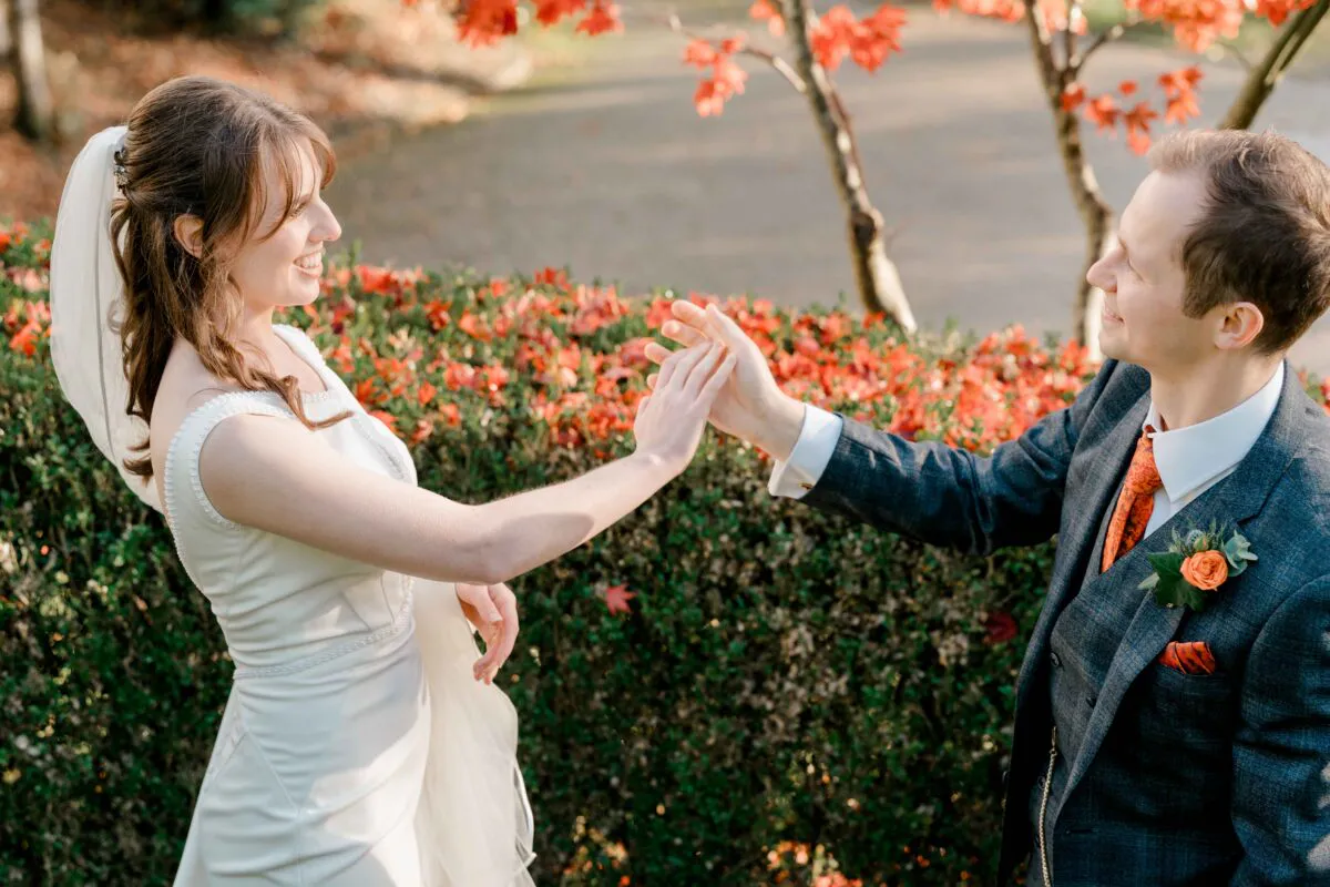 A bride and groom share a tender moment holding hands outdoors amidst vibrant autumn leaves and foliage during golden hour.