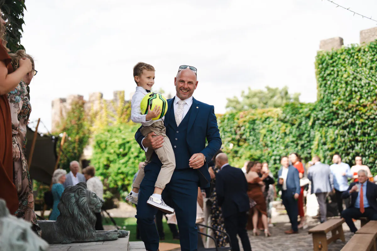 A smiling man in a navy three piece suit holding a young boy who is hugging a yellow and green football outdoors near an ivy-covered stone wall.