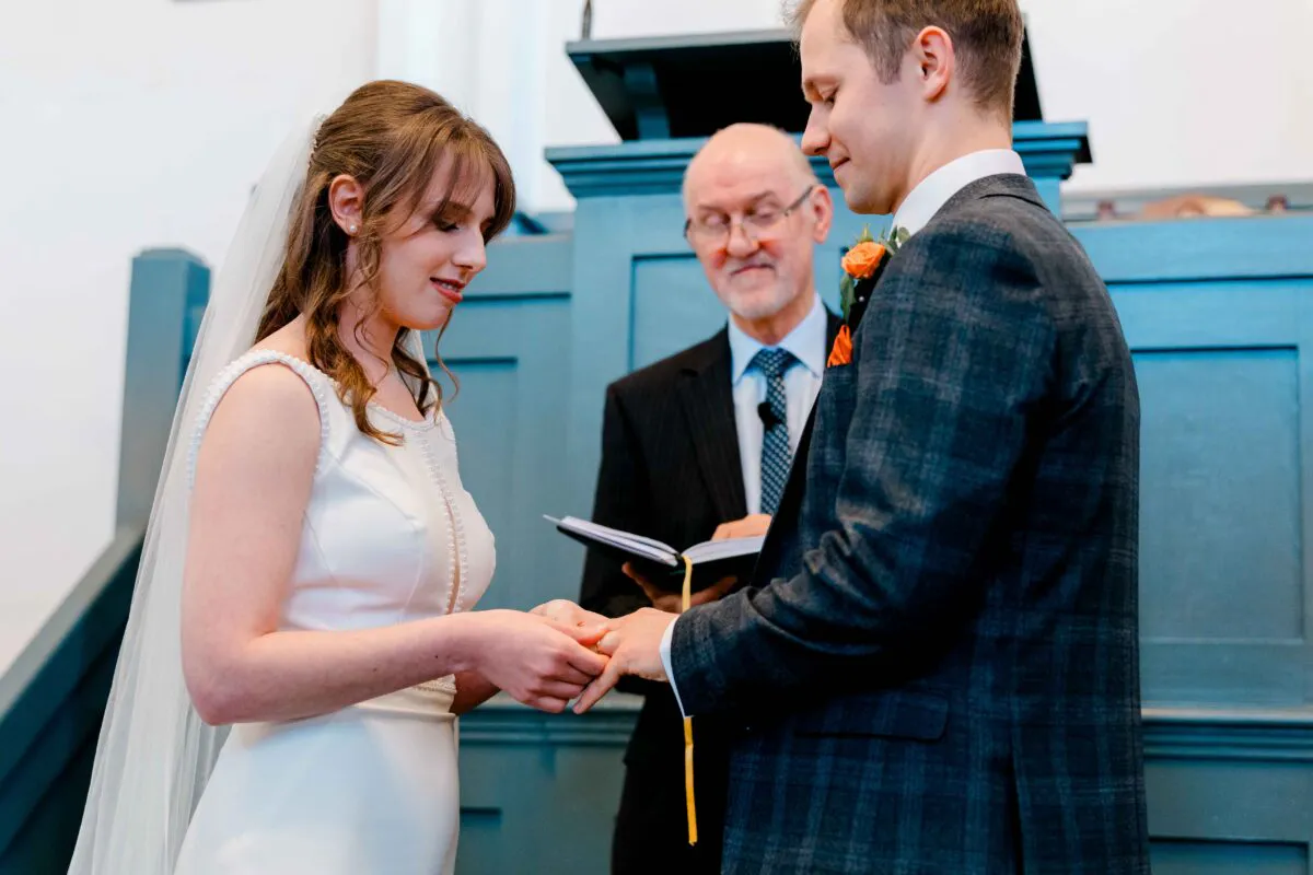 The bride gently places a wedding ring onto the groom's finger during a church ceremony, with the celebrant looking on closely.