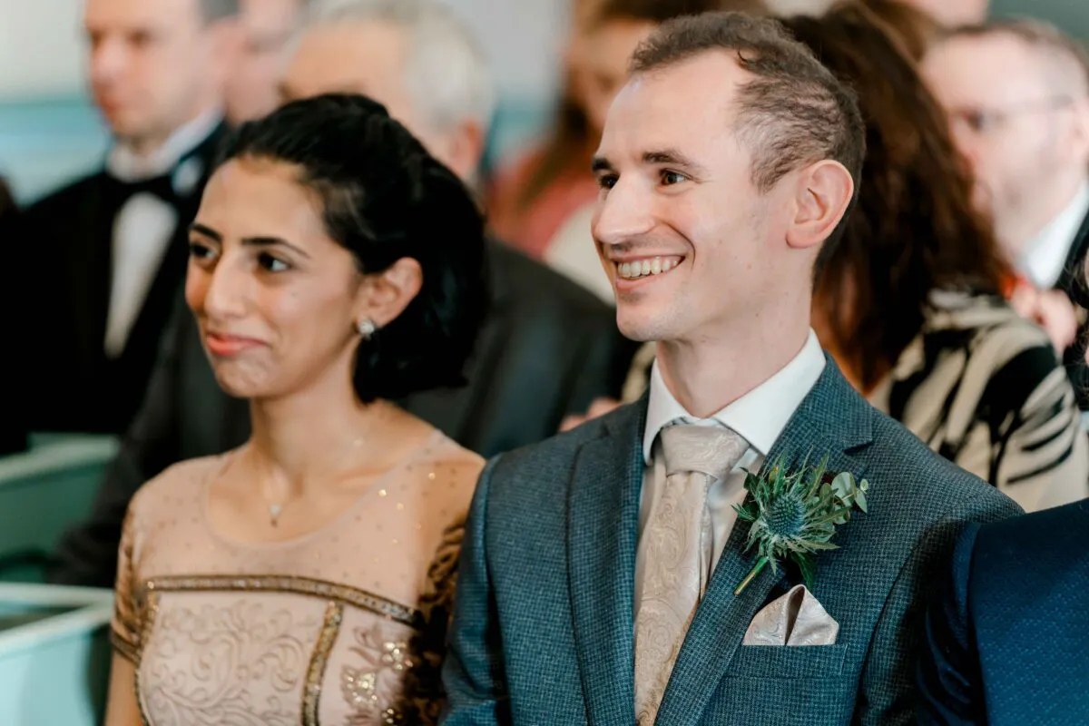 A smiling guest in a grey tweed suit with a thistle buttonhole sits alongside a guest in a decorative beaded dress during the wedding ceremony.