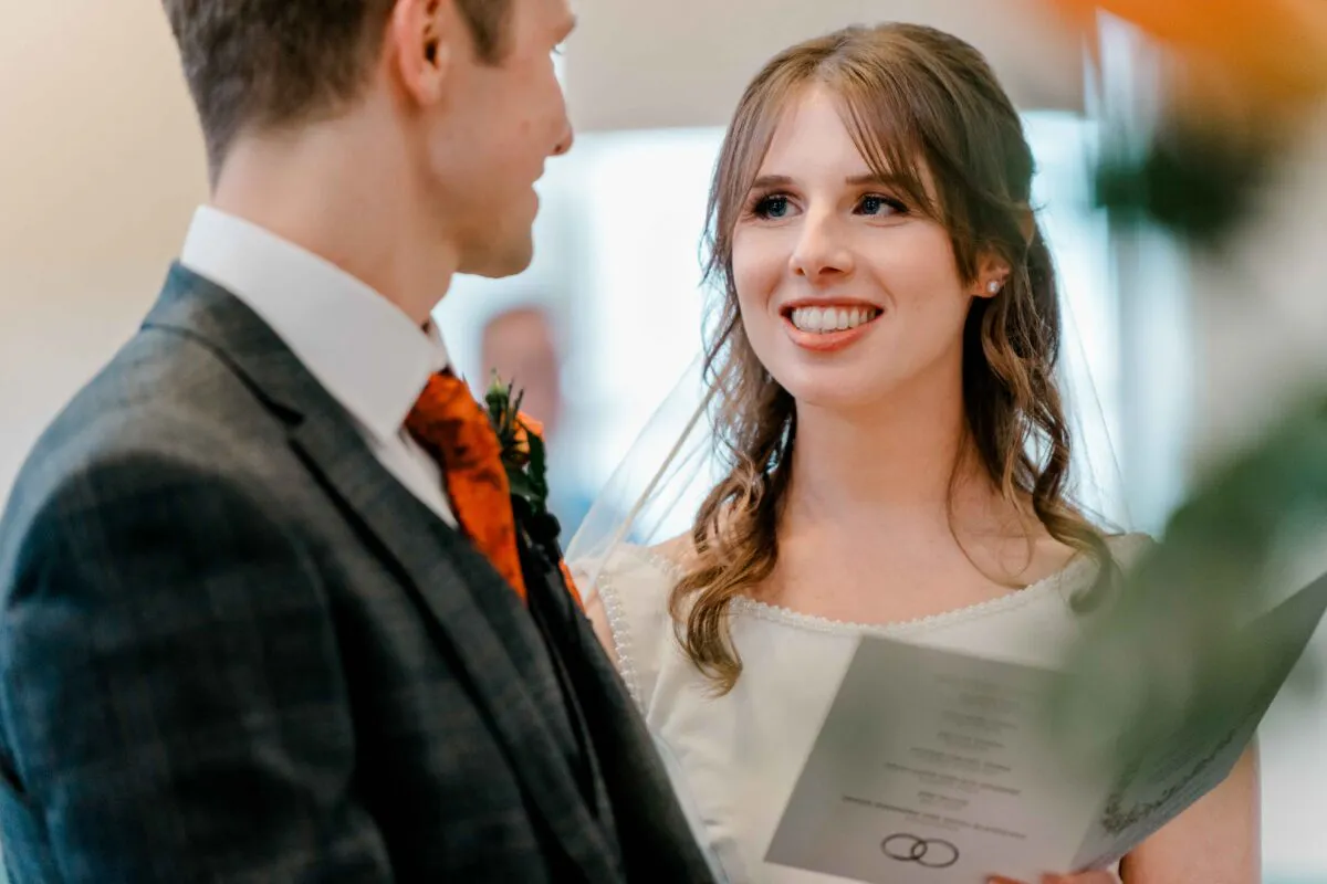 A smiling bride looks lovingly at her groom during their wedding ceremony while holding an order of service booklet.